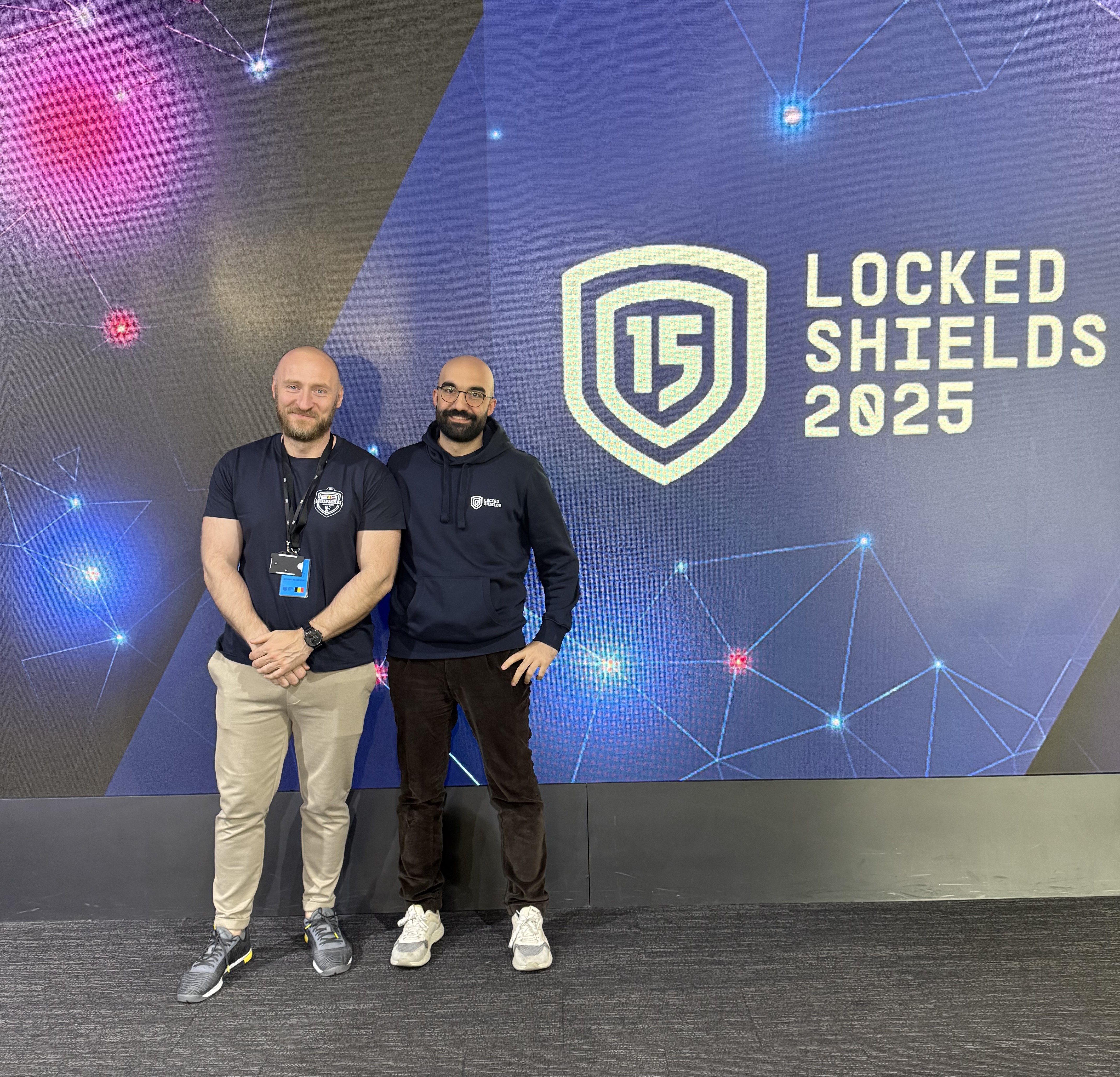 Two experts from Nexova in front of Locked Shields poster