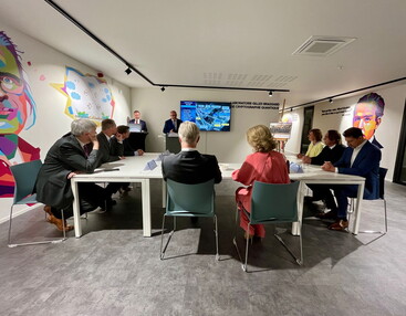 Nexova staff member standing in front of a large screen talking to Their Majesties, the King and Queen of Belgium who are seated alongside other people around a table